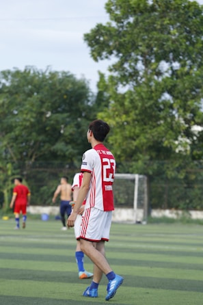A soccer player wearing a red and white jersey with the number 23 is walking on a green field. In the background, other players are visible, including one without a shirt. There are trees and a goalpost in the distance under a clear sky.