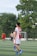 A soccer player wearing a red and white jersey with the number 23 is walking on a green field. In the background, other players are visible, including one without a shirt. There are trees and a goalpost in the distance under a clear sky.