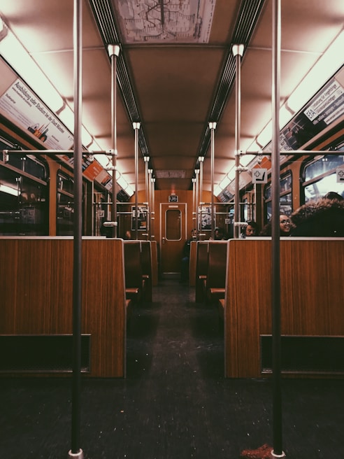 The interior of a vintage-style train carriage with wooden seats and metallic poles. Overhead advertisements and an old-fashioned ceiling map add to the retro ambiance. The benches are arranged in rows along the sides, and there are a few passengers visible.
