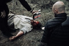 man in black bubble jacket facing man in white shirt lying on the ground while man in black bottoms holding digital time device