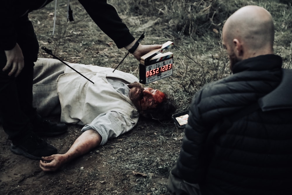 man in black bubble jacket facing man in white shirt lying on the ground while man in black bottoms holding digital time device