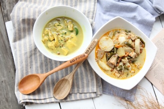 Two bowls of food with creamy dishes placed on a striped cloth, accompanied by wooden utensils. One bowl contains a yellow curry with pieces of chicken and garnishes, while the other features a mixed vegetable dish with a variety of ingredients in a creamy sauce. A pair of wooden utensils is displayed prominently on the cloth.