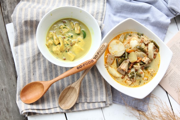 Two bowls of food with creamy dishes placed on a striped cloth, accompanied by wooden utensils. One bowl contains a yellow curry with pieces of chicken and garnishes, while the other features a mixed vegetable dish with a variety of ingredients in a creamy sauce. A pair of wooden utensils is displayed prominently on the cloth.