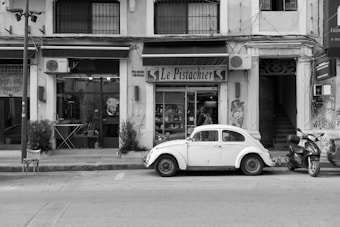 A vintage white car is parked on a city street in front of a shop named 'Le Pistachier.' The shop has a large display window and various items inside. A salon or caf&eacute; with a poster of a person inside is adjacent to it. There are two scooters parked nearby, along with plants and a chair on the sidewalk. The buildings have a slightly worn look, adorned with some graffiti and air conditioning units.