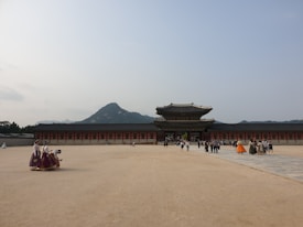 A historic palace stands in a wide-open courtyard under a clear sky. Visitors, some in traditional attire, walk around the area. A mountain rises in the background, providing a scenic backdrop.