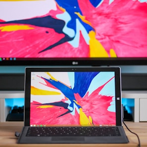 A laptop is positioned in front of a monitor, both displaying the same vibrant abstract artwork featuring bold splashes of red, blue, yellow, and black. The desk setup includes peripheral devices, and the color scheme of the artwork dominates the scene.