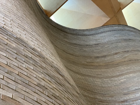 A curving wall made of light-colored bricks, with the bricks meticulously arranged to follow the contour. Above, a wooden ceiling structure with geometric patterns adds complexity to the architectural design.