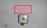 A cracked wall background with a red-bordered sign mounted above an emergency button. The sign reads 'IN CASE OF FIRE STOP AHU-1'.