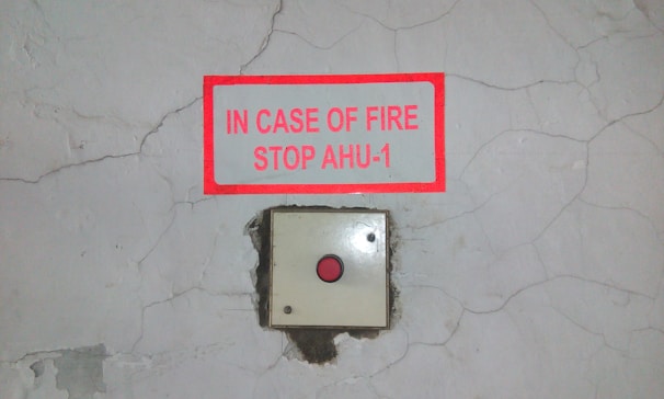 A cracked wall background with a red-bordered sign mounted above an emergency button. The sign reads 'IN CASE OF FIRE STOP AHU-1'.