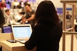 A person is standing at a podium, facing away from the camera with a laptop in front of them. The screen displays a document or presentation. In the background, several people are seated in an auditorium, some blurred and engaged in various activities, suggesting a conference or lecture setting.