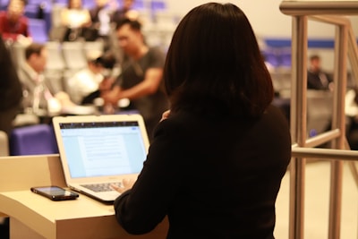 A person is standing at a podium, facing away from the camera with a laptop in front of them. The screen displays a document or presentation. In the background, several people are seated in an auditorium, some blurred and engaged in various activities, suggesting a conference or lecture setting.