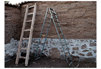 Two ladders, one wooden and leaning against a rough brick wall, and the other metallic and standing as a stepladder. The brick walls have patches of whitewash at the lower part near the ground.