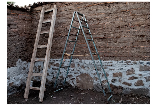 Two ladders, one wooden and leaning against a rough brick wall, and the other metallic and standing as a stepladder. The brick walls have patches of whitewash at the lower part near the ground.