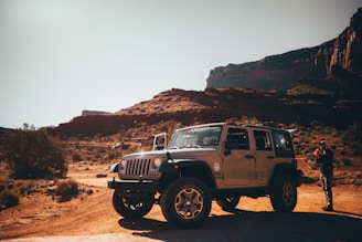 A rugged off-road vehicle is parked in a scenic desert landscape with rocky formations in the background. A person stands near the vehicle, holding something, possibly a camera or phone. The ground is covered with sparse vegetation, and the sunlight casts deep shadows.