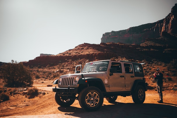 A rugged off-road vehicle is parked in a scenic desert landscape with rocky formations in the background. A person stands near the vehicle, holding something, possibly a camera or phone. The ground is covered with sparse vegetation, and the sunlight casts deep shadows.