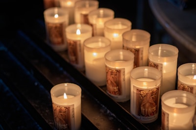 Several lit candles in glass holders are arranged on a dark surface. Each candle has a decorative image, likely of a religious figure, on the outside of the glass. The warm light from the flames illuminates the area softly, creating a peaceful and solemn atmosphere.