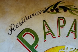 A rustic wall mural featuring the words 'Restaurant Papa'. The lettering is in vibrant shades, including green and red, with decorative olive branches above, adding an artistic and Mediterranean flair.