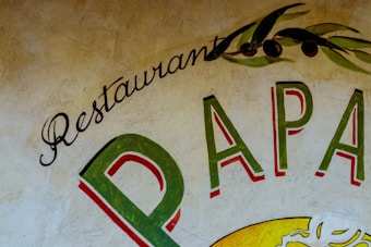 A rustic wall mural featuring the words 'Restaurant Papa'. The lettering is in vibrant shades, including green and red, with decorative olive branches above, adding an artistic and Mediterranean flair.