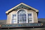 A building facade with a gabled roof and a decorative window featuring an intricate design. Below the window is a sign that reads 'Moccia's Train Stop' in elegant lettering.
