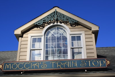 A building facade with a gabled roof and a decorative window featuring an intricate design. Below the window is a sign that reads 'Moccia's Train Stop' in elegant lettering.