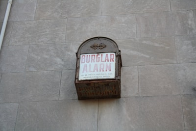 A vintage metal box attached to a stone wall with the words 'BURGLAR ALARM' written in bold red letters on a white background. Additional smaller text states 'O. B. McCLINTOCK CO. MINNEAPOLIS, MINN.'. The box appears to be weathered and rusting, suggesting it is an older alarm system.