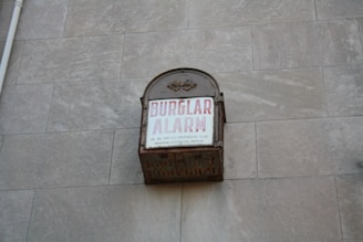 A vintage metal box attached to a stone wall with the words 'BURGLAR ALARM' written in bold red letters on a white background. Additional smaller text states 'O. B. McCLINTOCK CO. MINNEAPOLIS, MINN.'. The box appears to be weathered and rusting, suggesting it is an older alarm system.