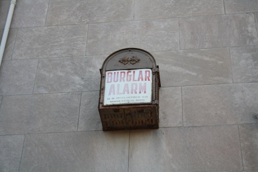 A vintage metal box attached to a stone wall with the words 'BURGLAR ALARM' written in bold red letters on a white background. Additional smaller text states 'O. B. McCLINTOCK CO. MINNEAPOLIS, MINN.'. The box appears to be weathered and rusting, suggesting it is an older alarm system.