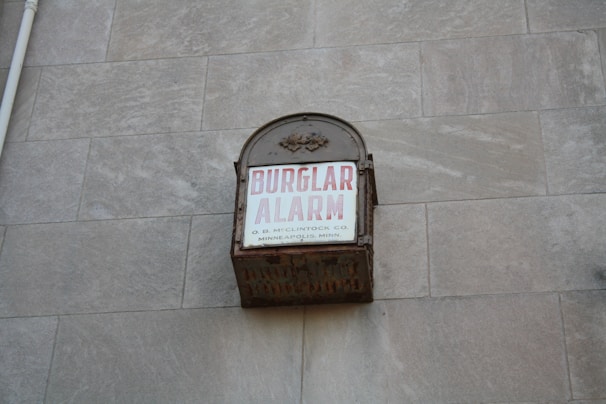 A vintage metal box attached to a stone wall with the words 'BURGLAR ALARM' written in bold red letters on a white background. Additional smaller text states 'O. B. McCLINTOCK CO. MINNEAPOLIS, MINN.'. The box appears to be weathered and rusting, suggesting it is an older alarm system.