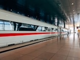 A sleek, modern high-speed train is stationed at a spacious, well-lit train station. The train is white with a distinctive red stripe running along its side, and the platform is marked with clear signage and reflective tiles. The overhead ceiling is dark and metallic, adding to the contemporary ambiance.
