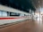 A sleek, modern high-speed train is stationed at a spacious, well-lit train station. The train is white with a distinctive red stripe running along its side, and the platform is marked with clear signage and reflective tiles. The overhead ceiling is dark and metallic, adding to the contemporary ambiance.