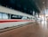 A sleek, modern high-speed train is stationed at a spacious, well-lit train station. The train is white with a distinctive red stripe running along its side, and the platform is marked with clear signage and reflective tiles. The overhead ceiling is dark and metallic, adding to the contemporary ambiance.