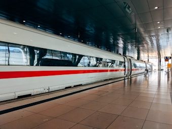A sleek, modern high-speed train is stationed at a spacious, well-lit train station. The train is white with a distinctive red stripe running along its side, and the platform is marked with clear signage and reflective tiles. The overhead ceiling is dark and metallic, adding to the contemporary ambiance.