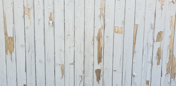 Weathered wooden boards with peeling white paint exposing patches of the natural wood underneath. Vertical alignment creates a pattern of alternating painted and bare surface areas.