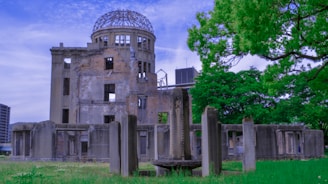 The image displays a ruined building surrounded by green grass and trees with a bright blue sky overhead. The structure's walls are mostly intact but show significant damage, with parts missing and exposed framework visible on the top. The foreground includes columns and remnants of what appears to be an old foundation or wall.