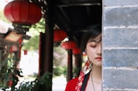A person in traditional clothing stands beside a gray brick wall, with their eyes gently closed. Red lanterns hang in the background, casting a vibrant, cultural atmosphere. Lush greenery is visible beyond the covered walkway, adding a touch of nature.