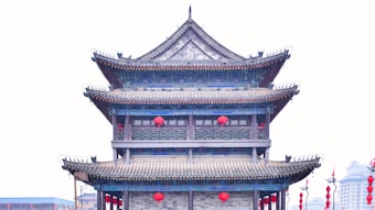 A traditional East Asian architectural structure featuring a multi-tiered pagoda style with intricate roof designs. The building's facade is adorned with red lanterns, enhancing the cultural aesthetic. The backdrop is a faint, overcast sky, emphasizing the structure's historical ambiance.