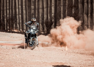 A motorcyclist wearing protective gear navigates a dirt track, kicking up a large cloud of dust behind the vehicle. The setting appears to be an off-road area, with ground markings and sparse vegetation visible.
