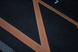 A dark asphalt surface with large orange diagonal lines creating geometric shapes. The texture of the road is visible, with some small specks and cracks.