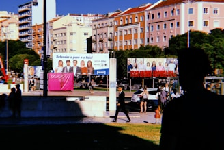A city street scene with colorful residential buildings in the background, and several large billboards featuring people and political messages. Silhouettes of pedestrians and a person in shadow are visible in the foreground. There's greenery around the area, and a few cars and people walking in the midground.