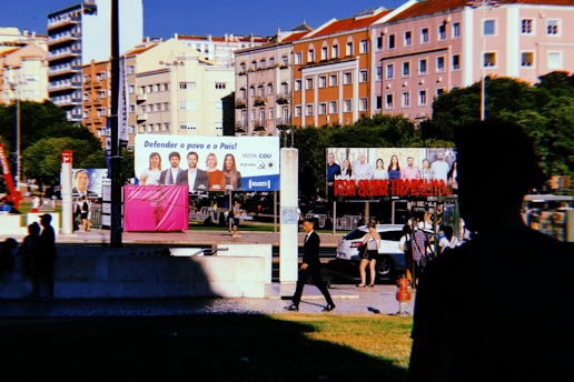 A city street scene with colorful residential buildings in the background, and several large billboards featuring people and political messages. Silhouettes of pedestrians and a person in shadow are visible in the foreground. There's greenery around the area, and a few cars and people walking in the midground.