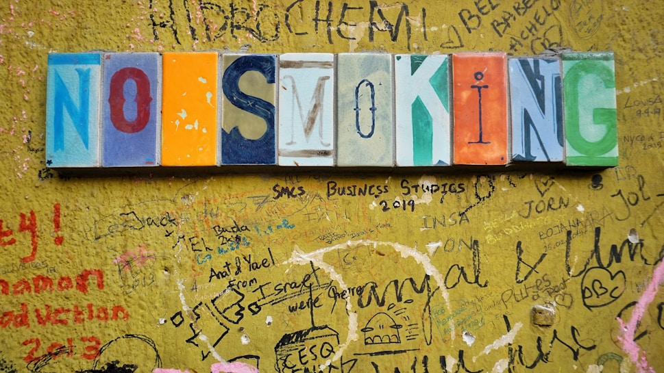 Colorful mosaic tiles spelling out 'NO SMOKING' are mounted on a vibrant yellow wall covered with various scribbles, drawings, and graffiti in different colors and handwriting styles.