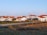 A serene rural village setting with several white houses featuring red-tiled roofs. The area is surrounded by fields and bordered by a dirt road. Power lines run across the foreground, indicating infrastructure. A few trees are visible among the houses.