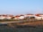 A serene rural village setting with several white houses featuring red-tiled roofs. The area is surrounded by fields and bordered by a dirt road. Power lines run across the foreground, indicating infrastructure. A few trees are visible among the houses.