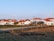A serene rural village setting with several white houses featuring red-tiled roofs. The area is surrounded by fields and bordered by a dirt road. Power lines run across the foreground, indicating infrastructure. A few trees are visible among the houses.