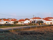 A serene rural village setting with several white houses featuring red-tiled roofs. The area is surrounded by fields and bordered by a dirt road. Power lines run across the foreground, indicating infrastructure. A few trees are visible among the houses.