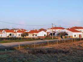 A serene rural village setting with several white houses featuring red-tiled roofs. The area is surrounded by fields and bordered by a dirt road. Power lines run across the foreground, indicating infrastructure. A few trees are visible among the houses.