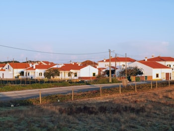 A serene rural village setting with several white houses featuring red-tiled roofs. The area is surrounded by fields and bordered by a dirt road. Power lines run across the foreground, indicating infrastructure. A few trees are visible among the houses.