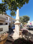 A stone monument stands in a quaint urban area, surrounded by colorful flowers and parked cars. The monument, with inscriptions likely indicating a historical significance, is flanked by two small basins. Trees with lush green leaves provide shade, and residential buildings with vibrant window trims line the street under a clear blue sky.