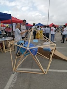 A group of people gathers at an outdoor event with tents in various colors. A geometric wooden structure is prominently displayed in the foreground, with two people interacting with it. The background shows other attendees and various activities taking place.