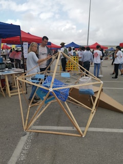 A group of people gathers at an outdoor event with tents in various colors. A geometric wooden structure is prominently displayed in the foreground, with two people interacting with it. The background shows other attendees and various activities taking place.